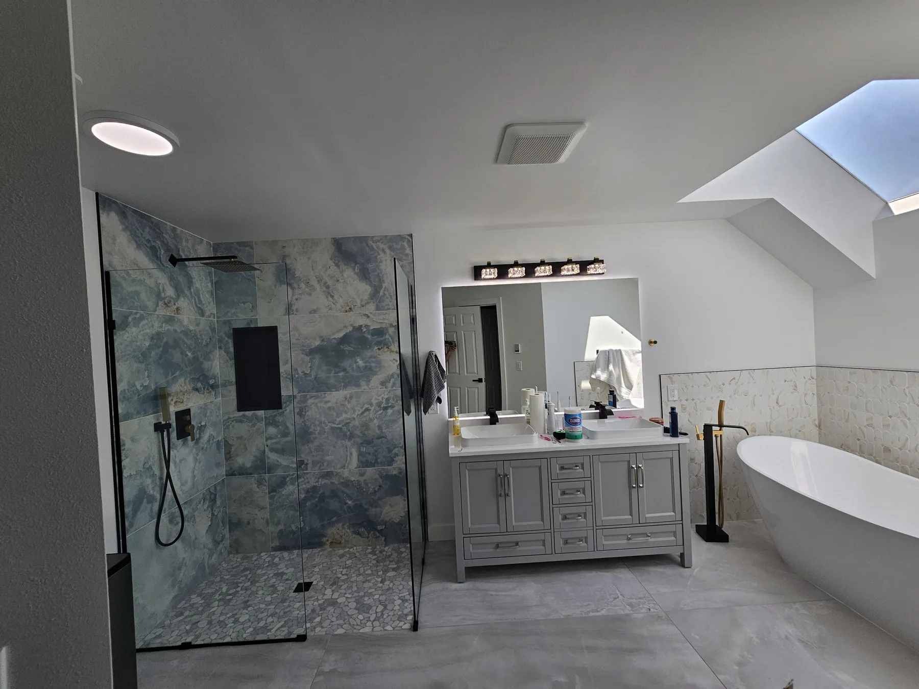 A finished primary bathroom with a blue marble walk-in shower, double vanity and freestanding tub under a skylight