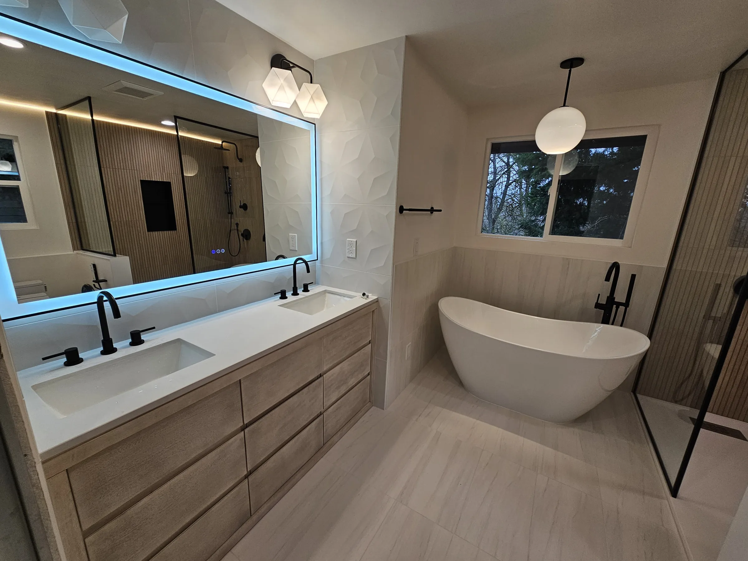 After — A bathroom in Juanita, Kirkland before and after renovation — an oak vanity and jetted tub replaced with a lit mirror, light oak double vanity and freestanding tub