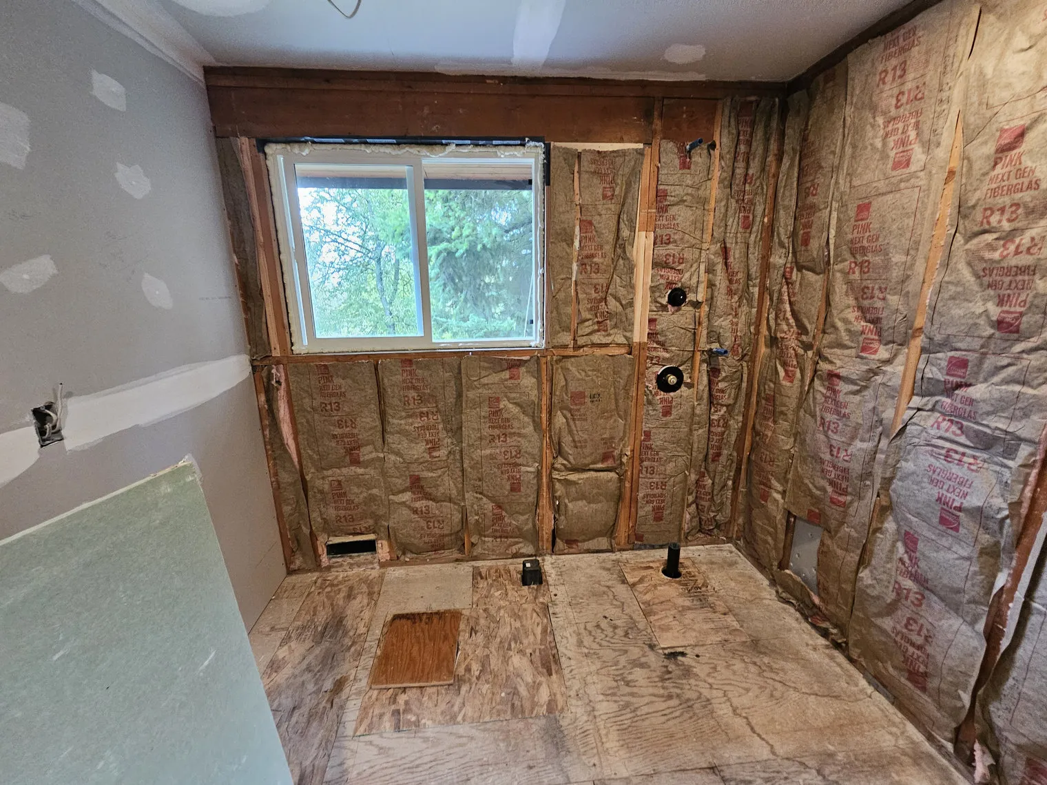 A bathroom stripped to studs and insulation, the window framing and wall cavities open before anything is rebuilt