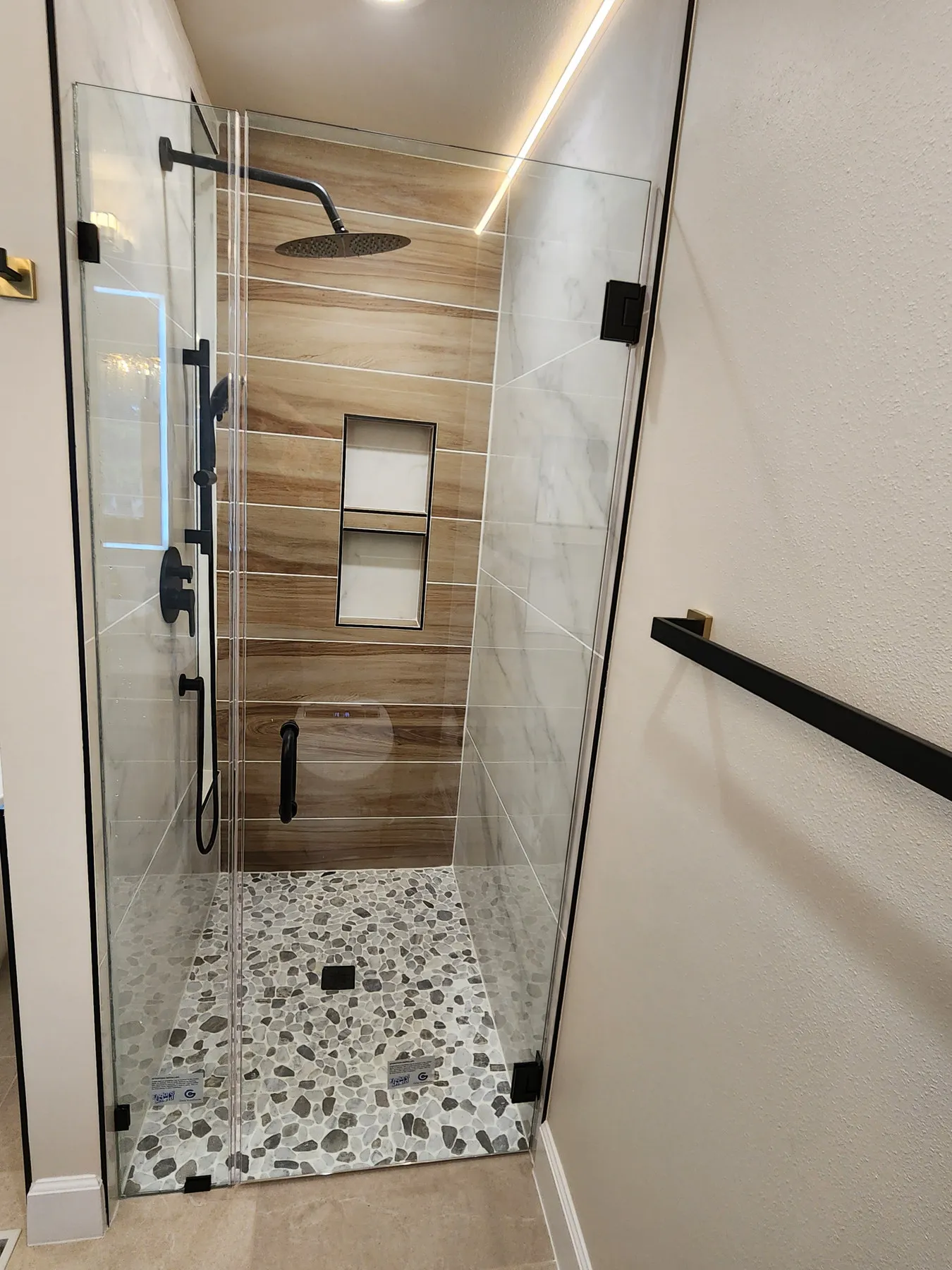 A walk-in glass shower with a wood feature wall and a pebble floor